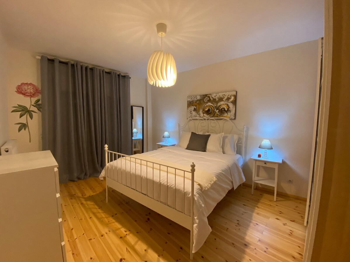 A well-lit bedroom features a spacious bed framed by soft bedding and pillows. Nightstands with lamps flank the bed, providing a gentle glow. A decorative wall painting and a large mirror enhance the ambiance, while elegant curtains create a sense of privacy.