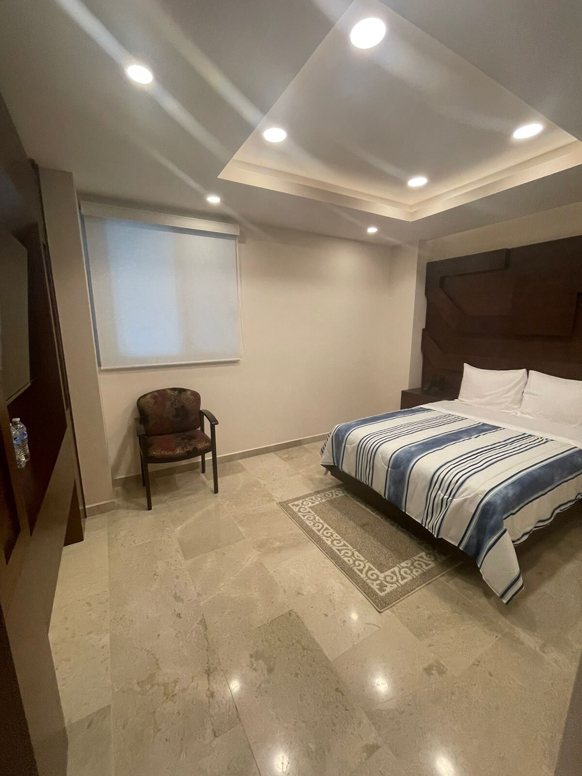 A well-appointed sleeping area features a spacious bed with striped bedding. Natural light enters through a window adorned with a simple roller shade. A patterned area rug adds texture to the marble floor, complemented by a comfortable chair positioned against the wall.