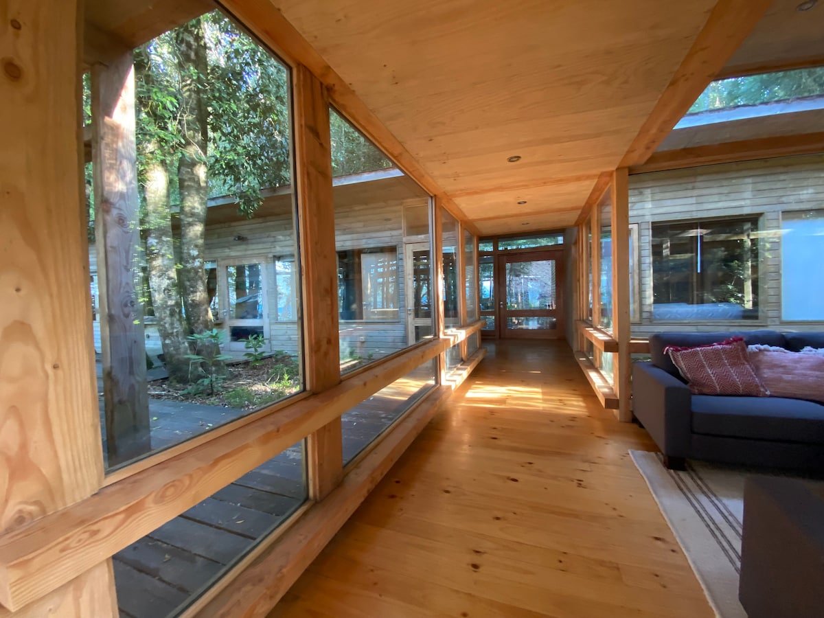 A corridor features extensive glass panels that invite natural light, connecting different sections of the house. Wooden walls and flooring provide a warm ambiance, while nearby trees create a calming atmosphere. A cozy seating area is visible at the end of the hall.