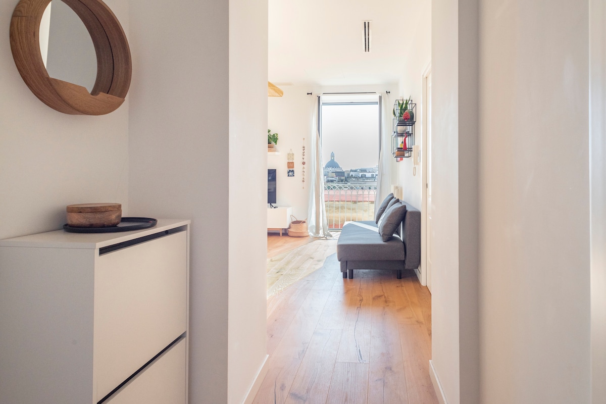 A bright hallway welcomes guests with a view into a living area featuring a comfortable gray sofa and a large window that frames an expansive outdoor view. Decorative elements include a circular mirror on the wall and a minimalist storage unit.