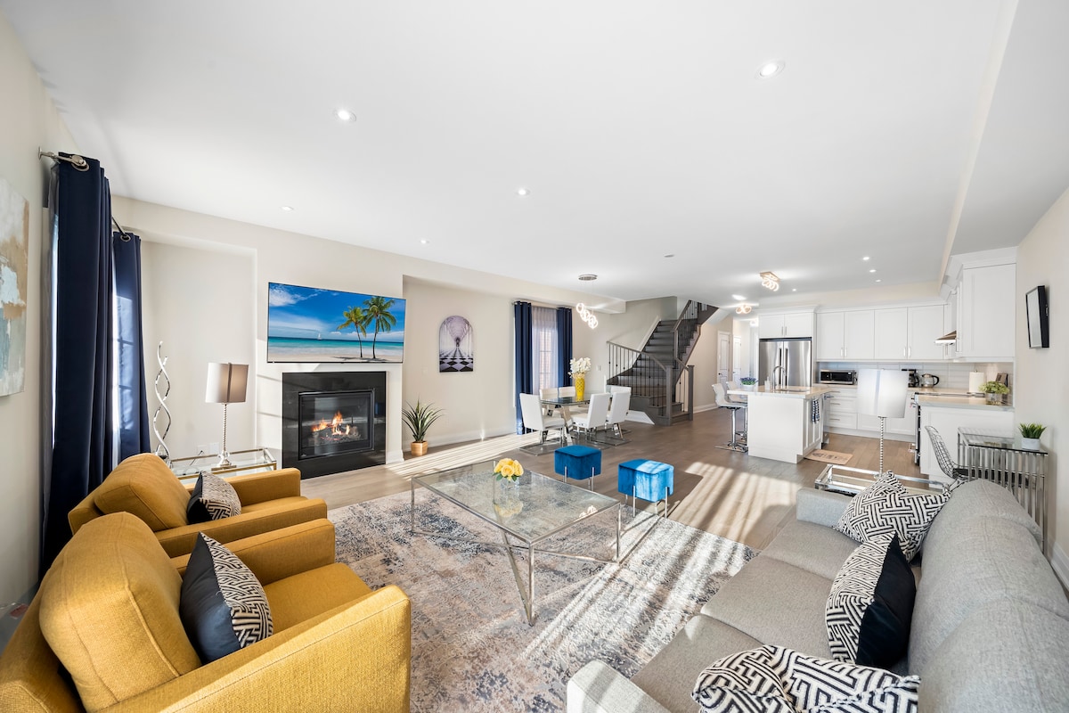 The spacious living area features a modern fireplace as the focal point, flanked by two comfortable sofas in muted tones. A large piece of artwork depicting a beach scene adorns the wall, while a bright area rug adds texture to the hardwood floor.