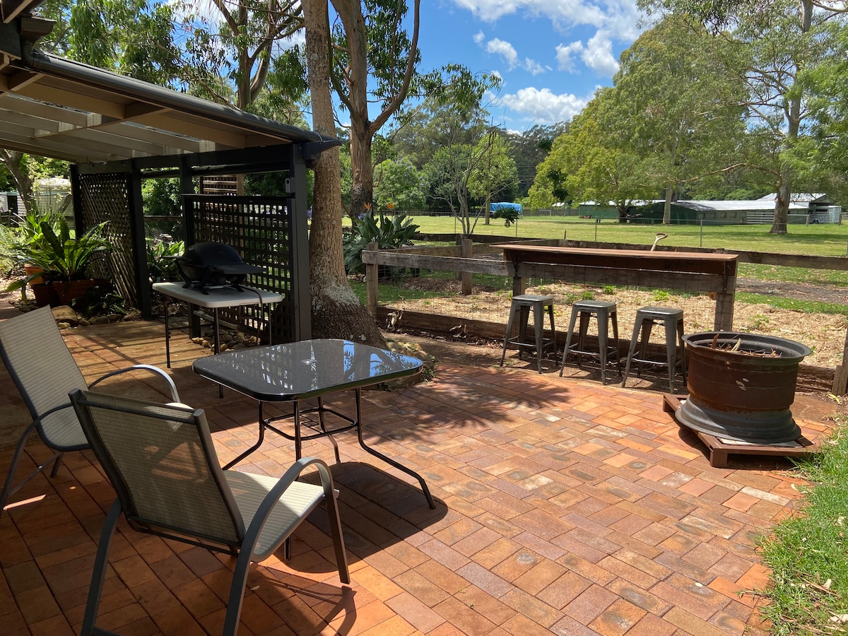 An outdoor seating area features two light-colored chairs and a glass-top table set on a terracotta tiled patio. A barbecue grill is positioned beneath an overhead patio cover, with a backdrop of lush greenery and open fields visible in the distance.