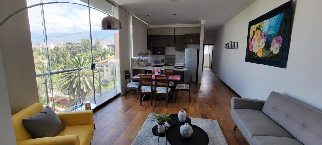 A bright living space features large windows offering views of palm trees and distant mountains. A spacious dining table is accompanied by six chairs, while a comfortable gray sofa and a vibrant yellow armchair provide seating. Decorative elements and a central coffee table enhance the modern design.