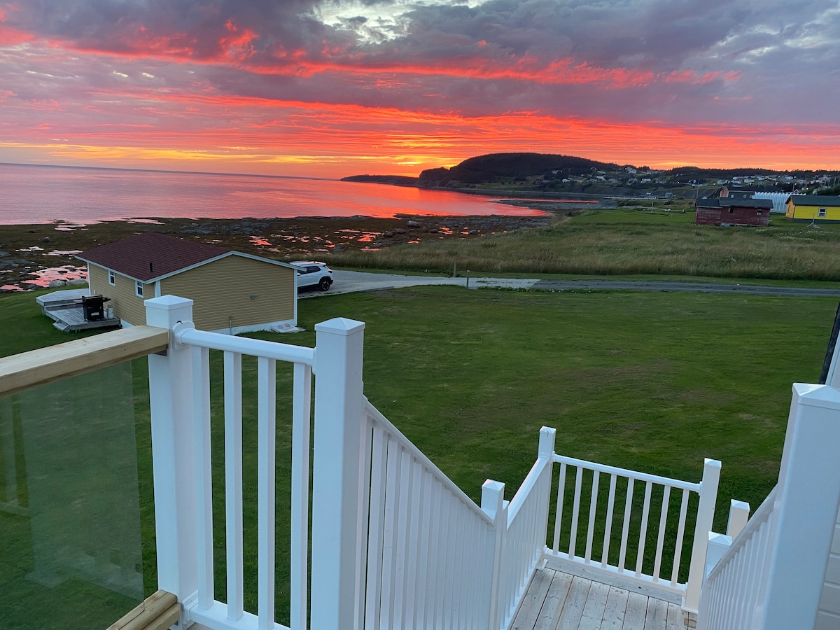 A stunning sunset is captured over the water, featuring vibrant hues of orange and pink in the sky. The view opens up to a calm ocean with reflections on the surface. A staircase with a white railing leads down from a deck, framed by lawn and a coastal landscape.