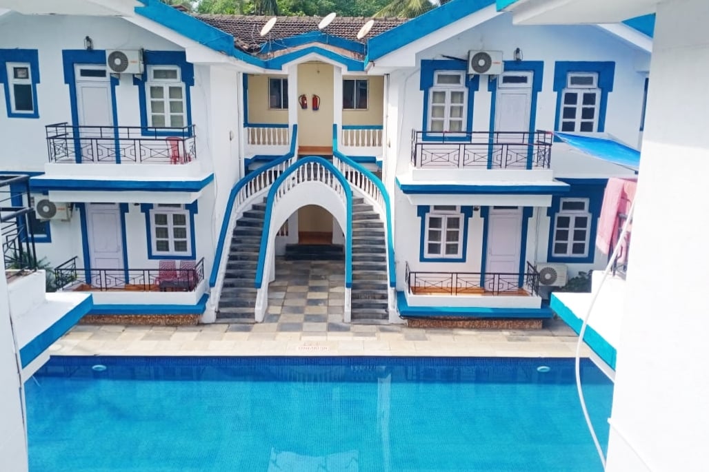 The image presents a gated complex featuring a clear swimming pool surrounded by elegantly designed blue and white buildings. A central staircase connects the two levels, enhancing accessibility. Air conditioning units are visible on the exterior walls, emphasizing comfort for guests.