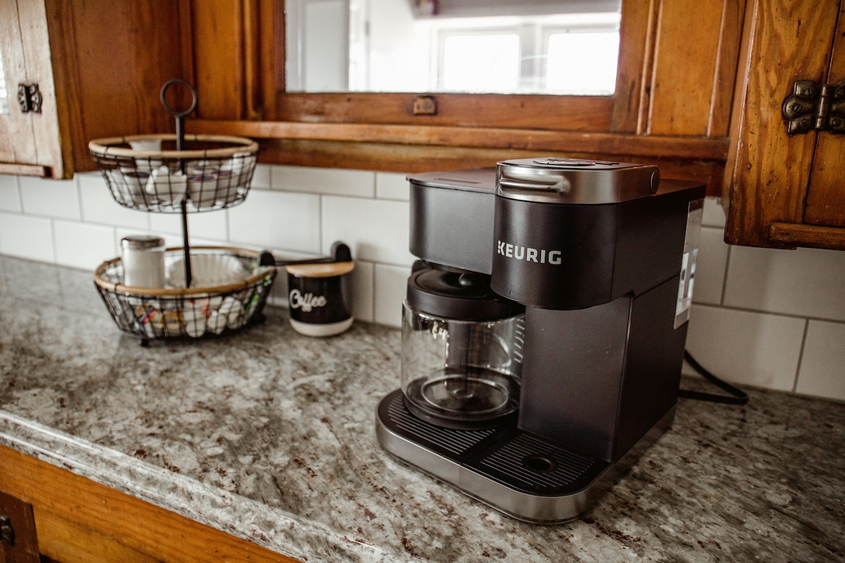 A well-equipped kitchen countertop showcases a Keurig coffee maker and a glass carafe. In the background, a wire basket holds various kitchen items. The space features light-colored tiles and polished wooden cabinetry, contributing to a functional and organized environment.