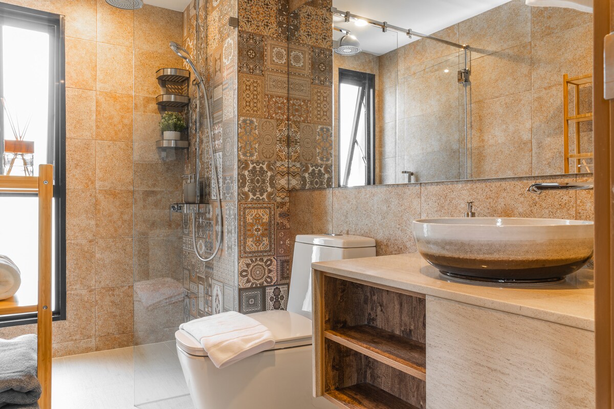 The bathroom features modern tile work with intricate patterns on the wall. A vessel sink sits atop a stone countertop, while a sleek shower area is equipped with a rain shower head. Natural light fills the space through large windows, complemented by minimalist storage solutions.