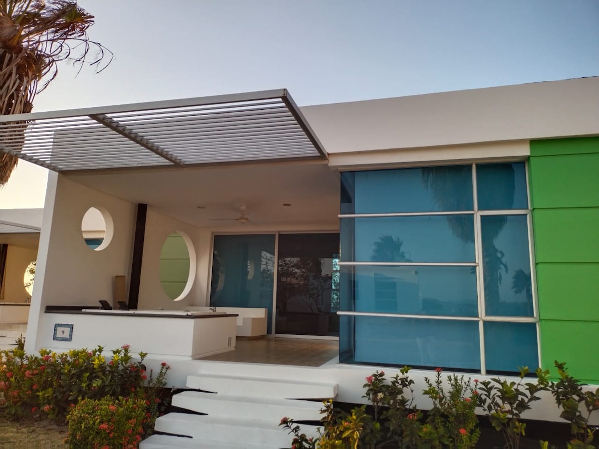 The exterior view of the cabin showcases a modern design with large glass panels and circular features. A covered terrace extends from the main living area, where a jacuzzi is visible. Lush greenery and colorful plants frame the entrance, enhancing the serene environment.