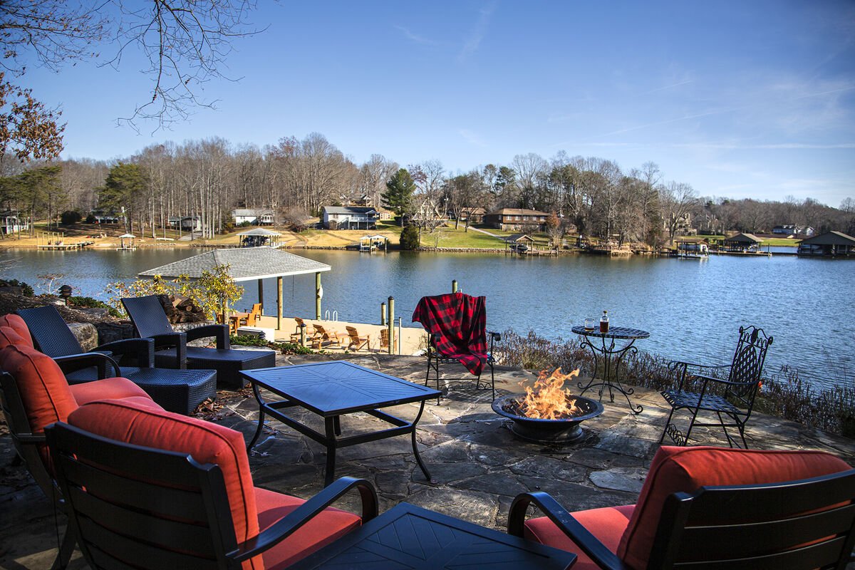 A lakeside patio is visible with several outdoor chairs arranged around a stone fire pit. A partial view of the lake is presented, showcasing calm waters and tree-covered banks. A covered area and additional seating can be seen across the water, creating a serene environment.