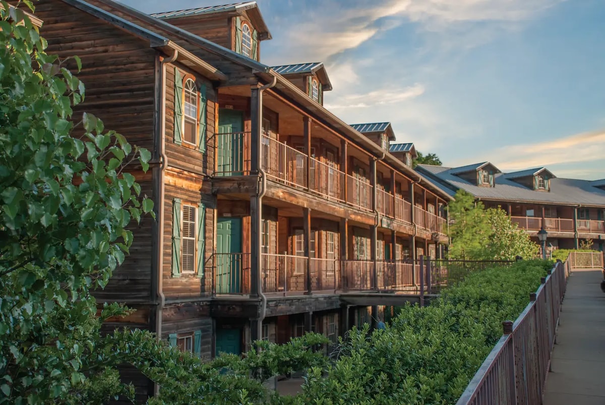 A multilevel wooden building features multiple balconies, adorned with green railings and vibrant teal doors. Lush greenery surrounds the structure, with a pathway leading towards the entrance, while soft clouds drift across the sky during sunset.