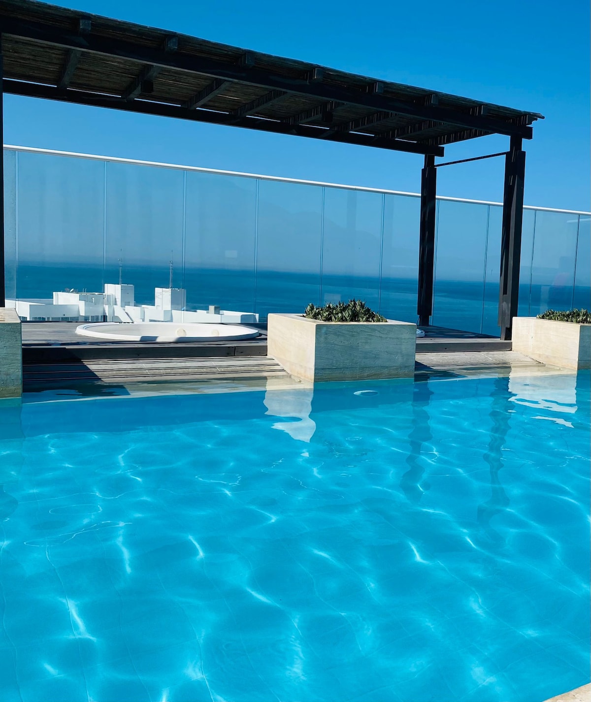 A refreshing rooftop pool is visible, surrounded by transparent glass fencing. The clear blue waters reflect the sunlight, creating a serene ambiance. In the background, endless ocean views can be seen, enhancing the tranquil setting.