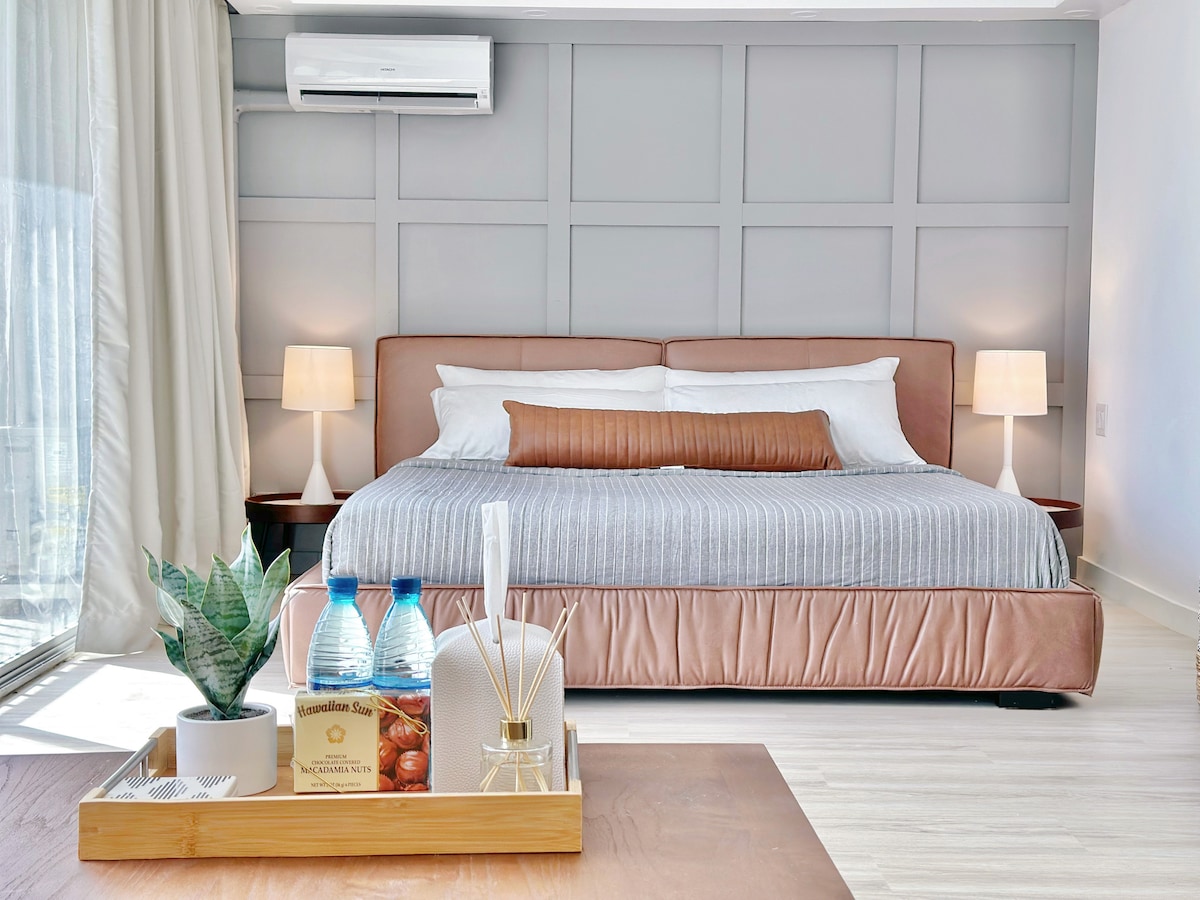 A spacious bedroom features a king size bed with a plush memory foam mattress, accented by soft bedding and decorative pillows. Two bedside lamps provide warm lighting, and a tray with water bottles and a fragrance diffuser is placed on a wooden table. Large windows allow natural light to enter.