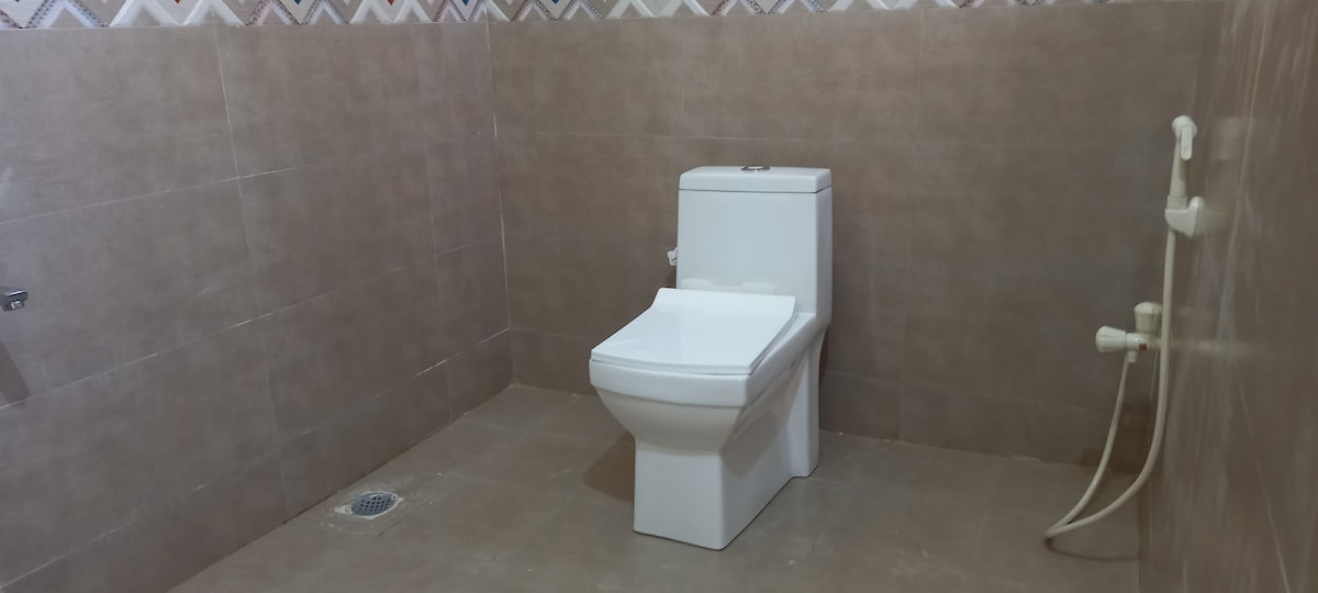 The bathroom features a modern, white toilet positioned against a neutral tiled wall. The space is kept minimalistic, with clean lines and a tiled floor. A water inlet and outlet are visible on one side, providing functionality without clutter.