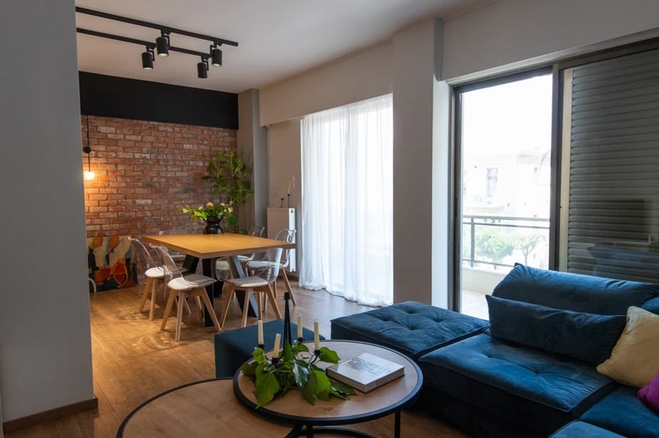 A modern living area features a comfortable blue sectional sofa and a round coffee table with greenery. Natural light fills the space through large glass doors, while an inviting dining area with a wooden table and stylish chairs is situated nearby against a brick accent wall.