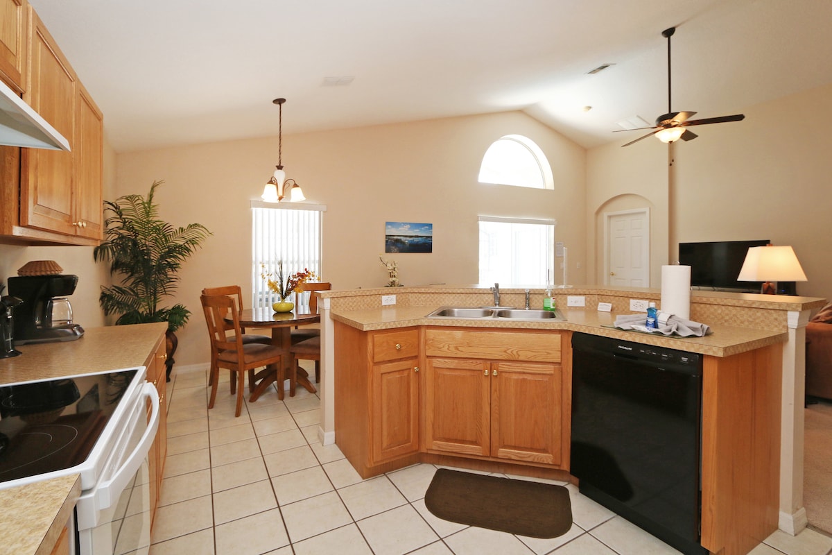 The kitchen features wooden cabinetry, a dark dishwasher, and modern appliances. A breakfast bar offers seating along the counter. Natural light enters through an arched window above the kitchen sink, while a dining table with wooden chairs is visible in the adjacent area.