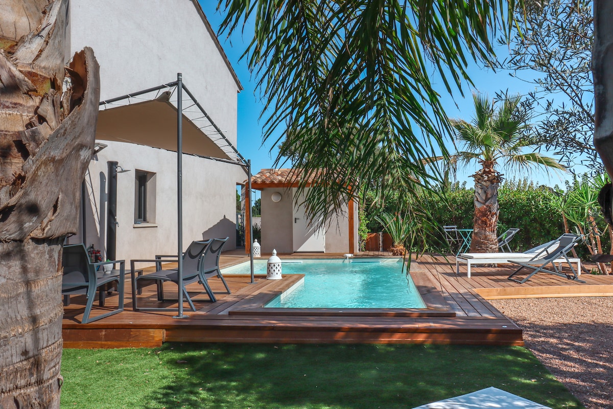 A private pool is surrounded by wooden decking, complemented by lounge chairs and outdoor seating. Lush greenery and palm trees frame the scene, providing a relaxing atmosphere. The modern villa stands in the background, showcasing large windows and an inviting entrance.