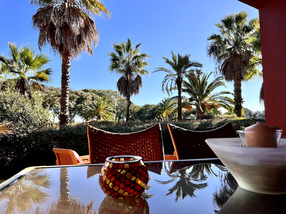 An outdoor terrace is depicted, featuring a glass table reflecting the surrounding space. Colorful palm trees provide a lush backdrop, while woven chairs add a comforting touch. A decorative candle holder and a bowl are placed on the table, enhancing the inviting atmosphere.