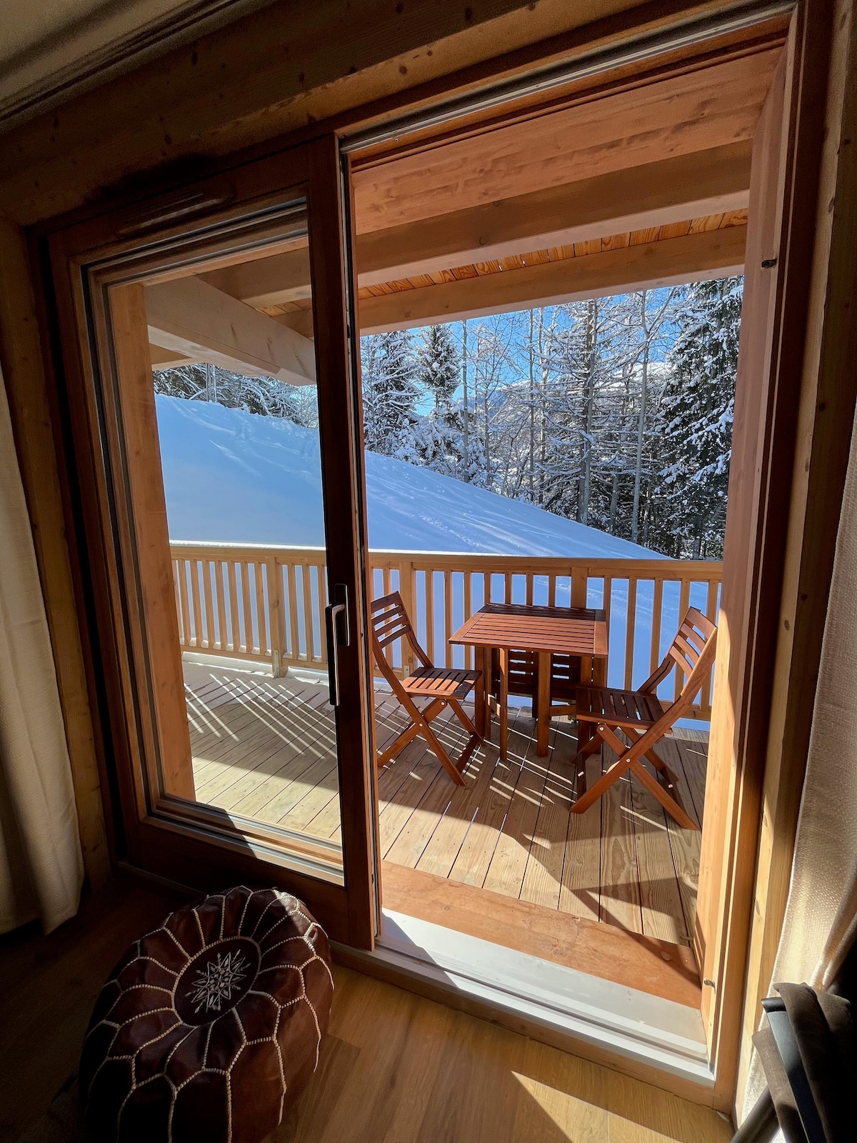 A wooden balcony is visible through large glass doors, featuring a small table and two folding chairs. Snow-covered trees can be seen in the background, and the serene atmosphere is enhanced by bright natural light.