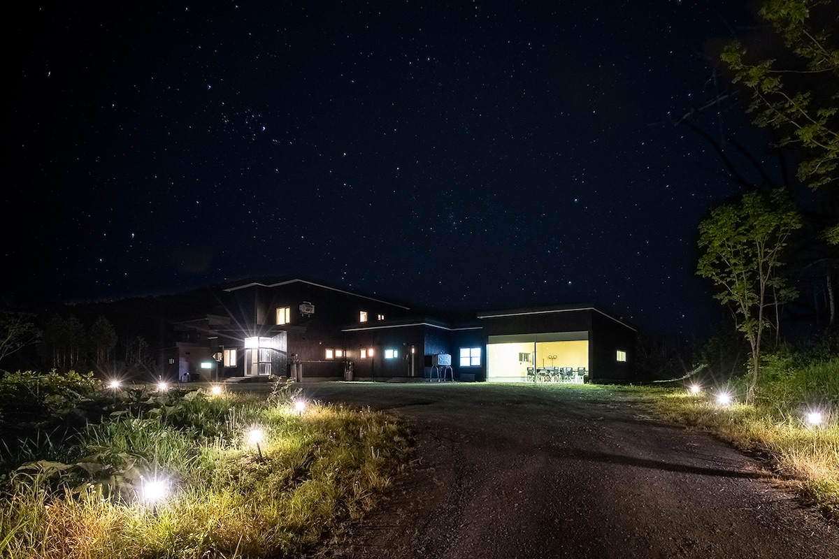 The exterior of the property is illuminated at night, showcasing a two-story building surrounded by a natural landscape. Soft lights line the pathway, and the starry sky above adds to the serene atmosphere. Windows reflect warm light from inside, suggesting a welcoming space.