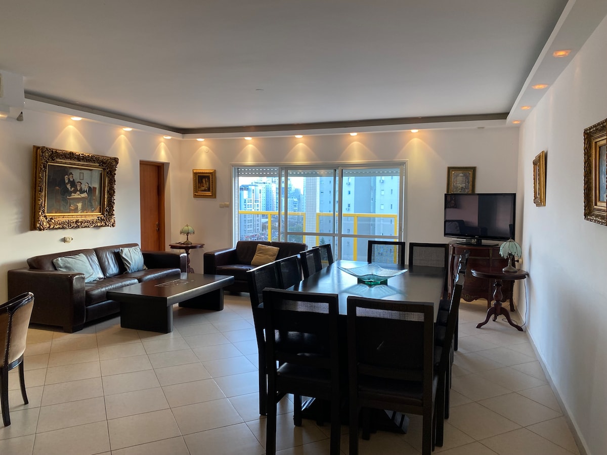 A spacious living area features elegant furnishings, including a dark leather sectional sofa and a wooden dining table with multiple chairs. Large windows provide ample natural light, and framed art adorns the walls, contrasting with the neutral tones of the space.