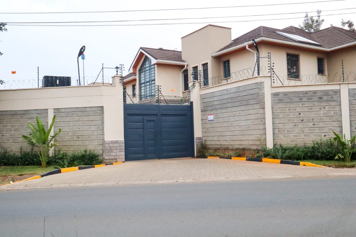 The property exterior features a modern design with a tall, gated entrance adorned by vertical slats and a concrete wall. Lush greenery borders the driveway, which is paved with stone, offering an inviting approach to the home. Security elements are visible atop the perimeter fence.