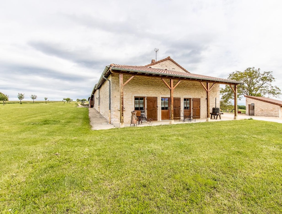 A historic agricultural building from the 18th century is prominently situated on a verdant plateau. The structure features wooden shutters and an inviting porch area. Surrounding green fields enhance the serene countryside atmosphere, with trees dotting the landscape in the background.