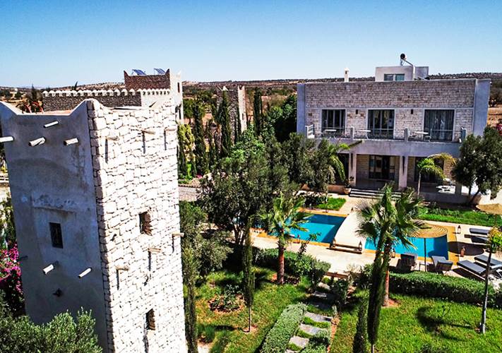An aerial view showcases a modern villa alongside a historical stone tower. The villa features expansive glass windows and is surrounded by lush greenery, including palm trees and flowering plants. A clear pool is positioned in front, inviting relaxation and outdoor enjoyment.