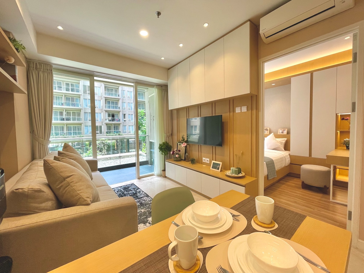 A spacious living area features a comfortable sofa facing a wall-mounted TV, with natural light streaming through glass doors that open to a balcony. A neatly set dining table with white dishware is positioned nearby, complementing the modern cabinetry and decor.