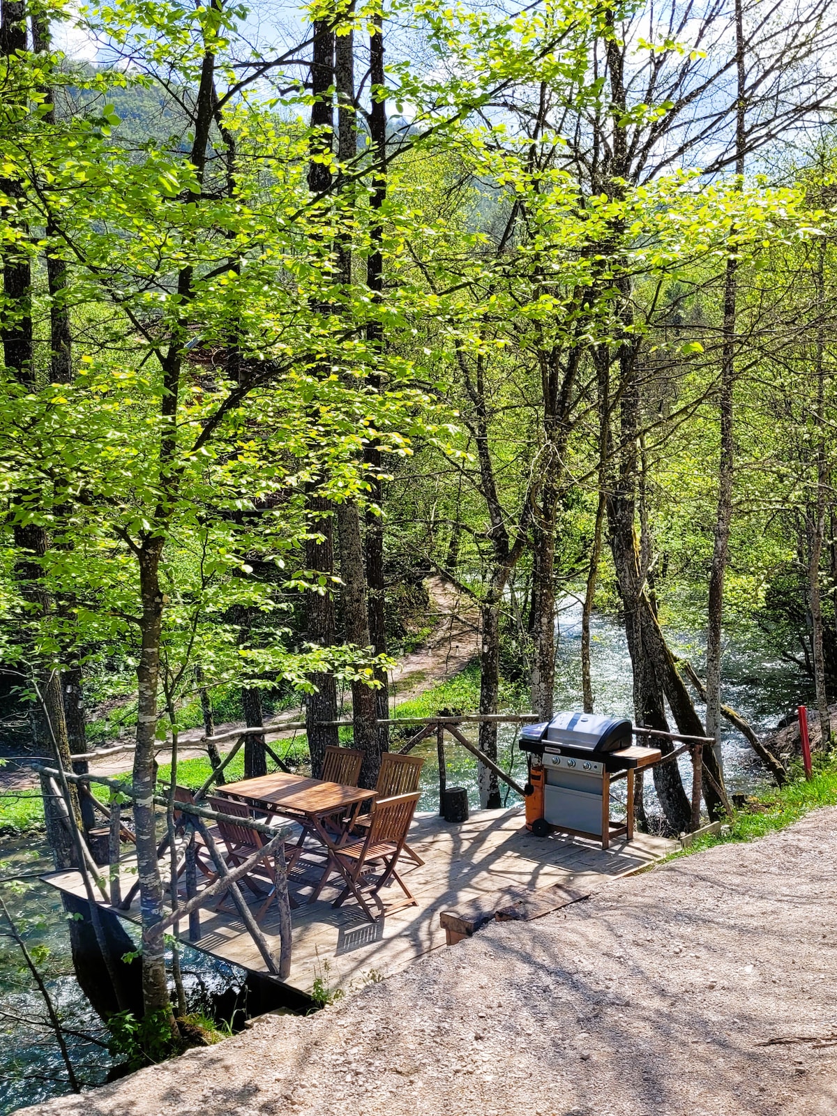 A wooden deck is positioned among lush green trees, with a dining table and chairs arranged nearby. A grill is available for outdoor cooking, and the serene river flows gently beside the space, enhancing the natural surroundings.
