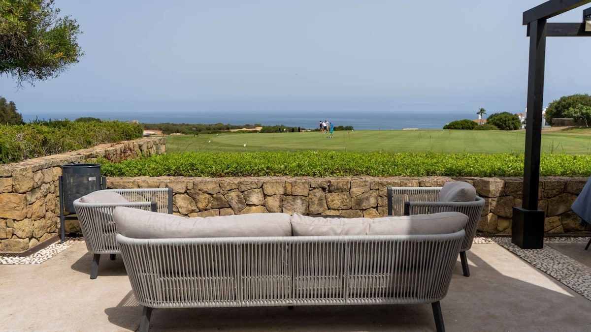 A comfortable outdoor seating area is positioned against a stone wall, offering unobstructed views of the golf course and the distant sea. The arrangement features a modern couch with soft cushions, surrounded by lush greenery and a clear blue sky.