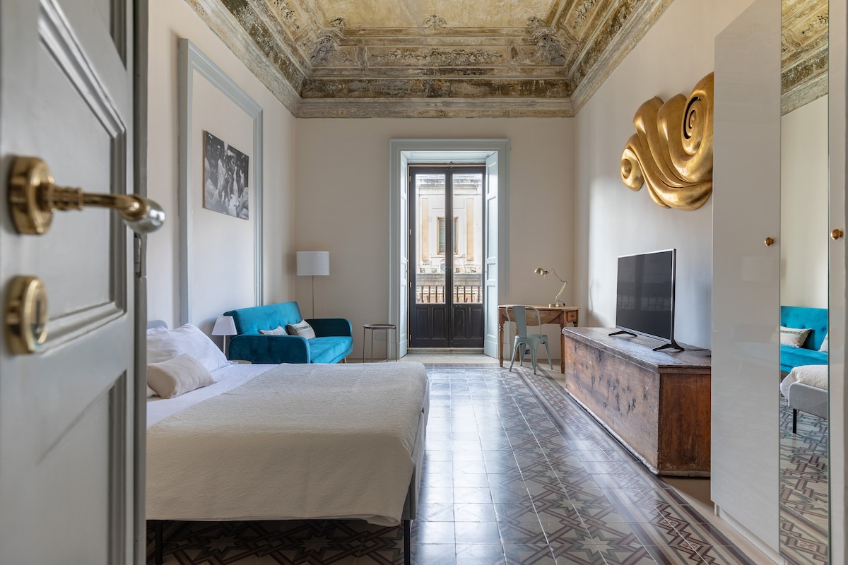 A spacious room features high, decorated ceilings and large windows that open onto a balcony. A comfortable bed is positioned next to a blue sofa, while a TV is located on a wooden sideboard. A mirror and lamp add functionality to the space.