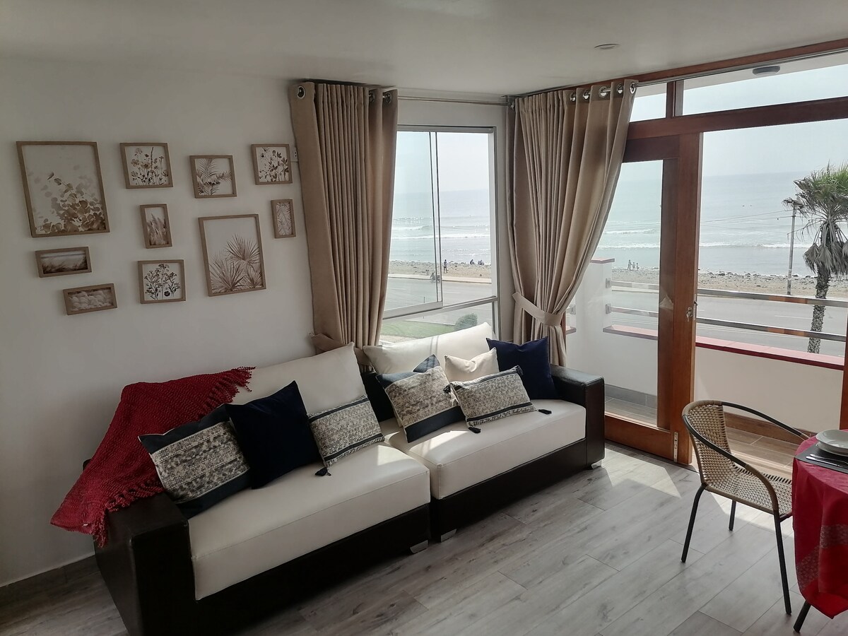 A comfortable seating area is presented with a white sofa adorned with navy blue and patterned cushions. Large windows provide natural light and showcase ocean views, while light curtains softly frame the space. A small round table and chairs complete the inviting ambiance.