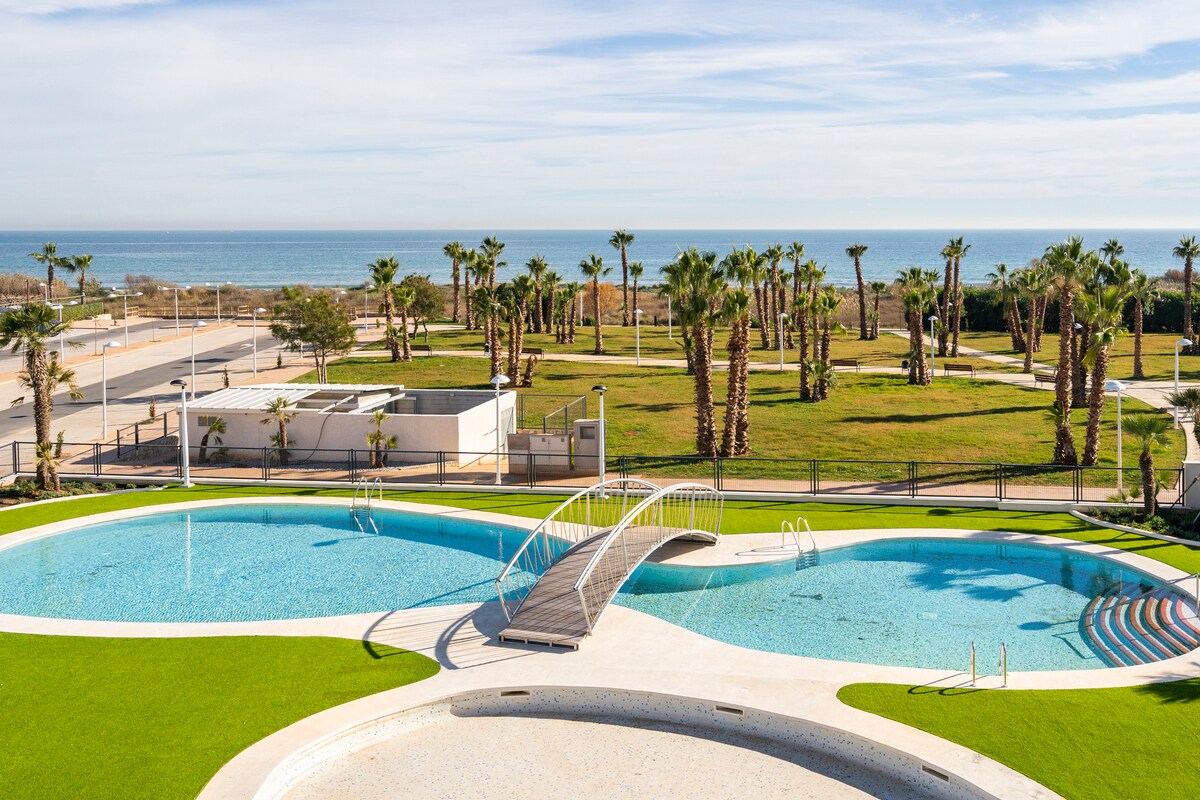 A view of a spacious outdoor pool area, featuring a circular pool with a small bridge, surrounded by manicured grass and palm trees. The ocean can be seen in the background, enhancing the scenic landscape.