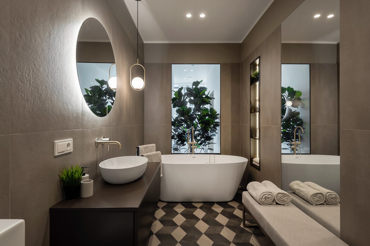 A modern bathroom is highlighted by a freestanding tub and a large, round mirror. A sleek vessel sink and contemporary fixtures are present. Plants are visible through large windows, providing a touch of nature. Textured flooring adds visual interest to the space.