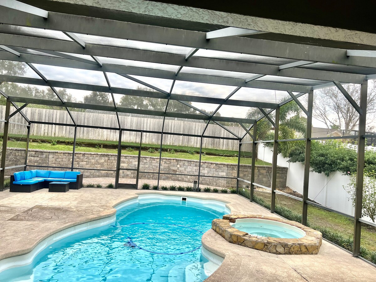 A spacious pool area features a sparkling blue pool beneath a screened enclosure. A circular hot tub is situated adjacent to the pool, surrounded by a stone border. Ample outdoor seating is provided on a blue couch, adding to the inviting atmosphere.