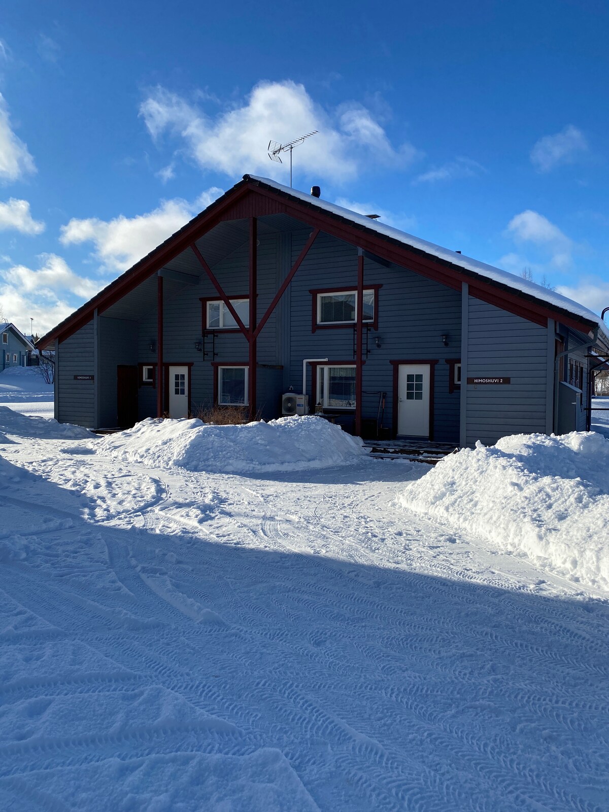 A two-story cabin is showcased against a snowy landscape, featuring a light blue exterior with red accents on beams and doors. The entrance is flanked by snow drifts, and clear skies are visible above, contributing to a serene winter scene.