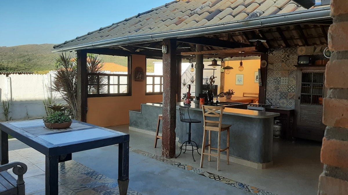An open kitchen area features a stone countertop with bar stools and hanging light fixtures. A wooden table with a potted plant is visible, and large windows allow natural light into the space. The rustic elements and patterned tiles enhance the inviting ambiance.