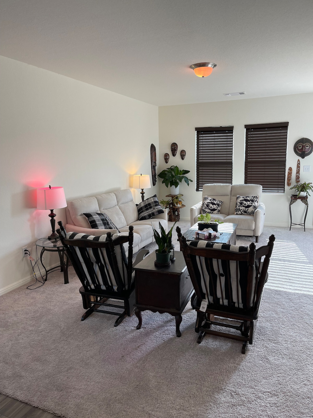 A light-filled living area features a cozy seating arrangement, including two plush armchairs and a comfortable sofa. Decorative plants add greenery, while art pieces and masks adorn the walls. Natural light streams through the window treatments, creating a calm and inviting atmosphere.