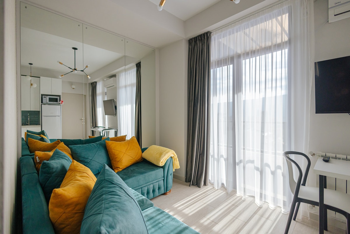 A cozy living area features a teal sofa adorned with yellow throw pillows. Light filters through sheer curtains, illuminating a modern design. A small dining table and chairs are visible, with a wall-mounted TV in the corner. An open kitchen area can be seen in the background.