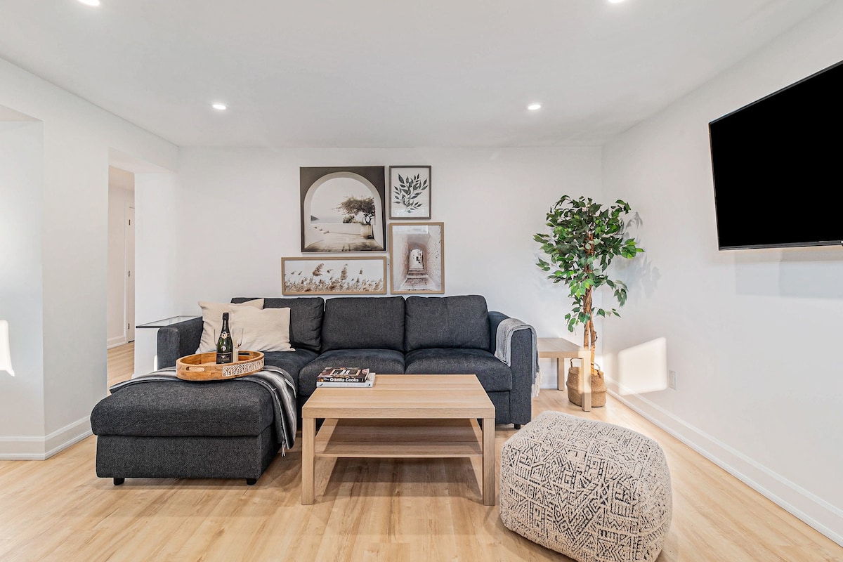 A spacious living area features a comfortable sectional sofa adorned with a soft throw blanket. A wooden coffee table is centered on the light wood flooring, accompanied by a decorative pouf. A large TV is mounted on the wall beside framed artwork and a potted plant.