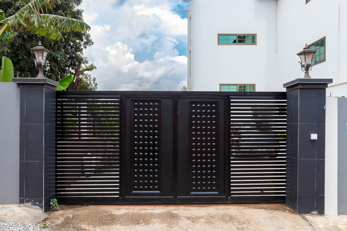 A modern, black gated entrance stands as the entrance to the property, featuring horizontal metal slats and decorative details. Flanking the gate are two stylish lampposts. The driveway is visible, surrounded by a tidy landscape with greenery in the background.