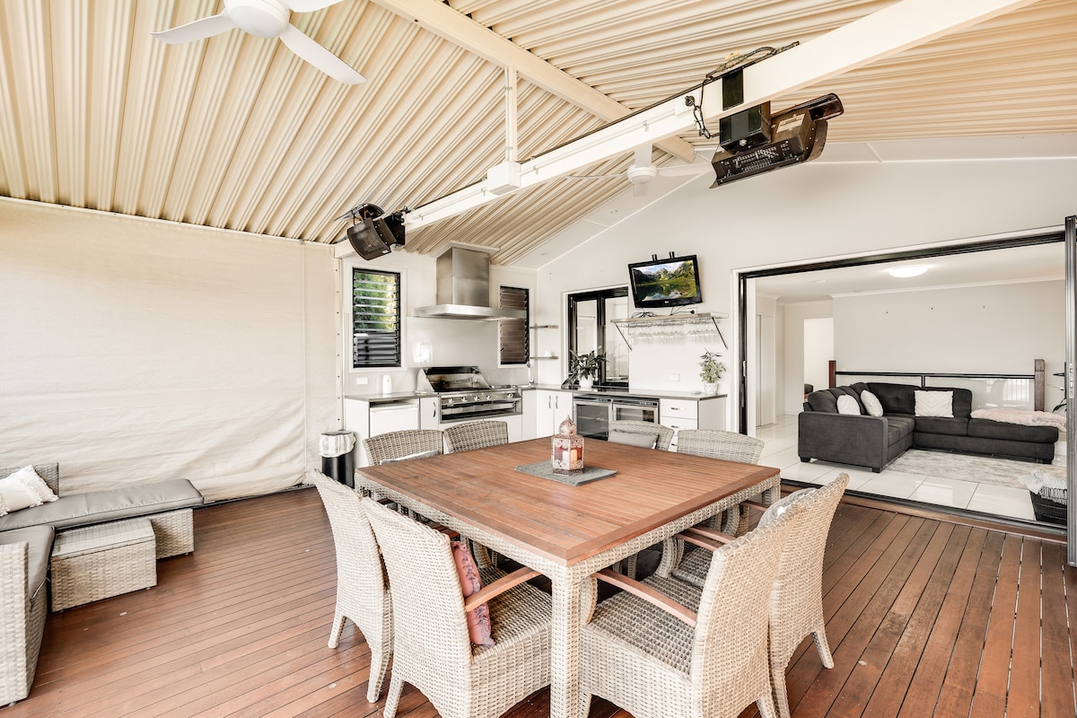 A spacious outdoor entertainment area is featured with a large wooden table surrounded by wicker chairs. An outdoor kitchen with modern appliances is visible, along with a wall-mounted TV. The area is shaded by a covered roof, providing comfort in various weather conditions.