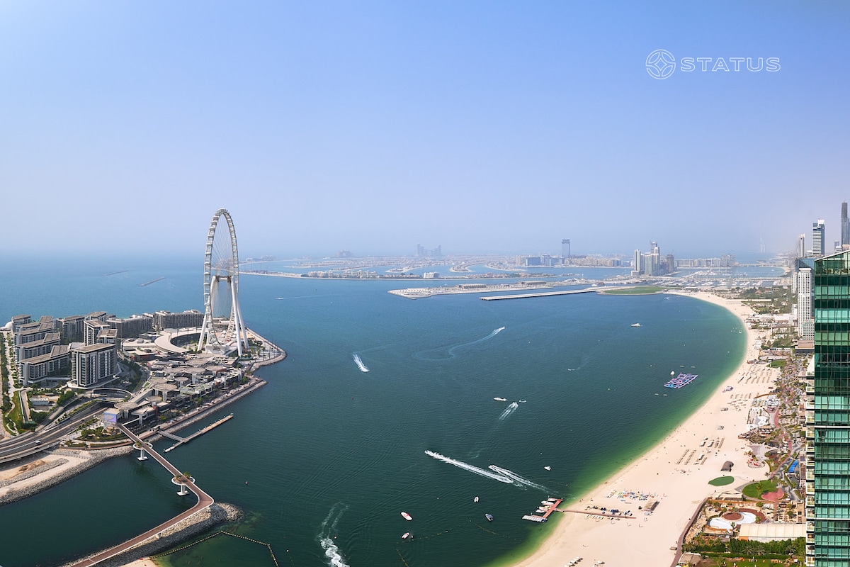 An expansive view of the coastline features the iconic Ain Dubai Ferris wheel in the foreground. The sandy beach stretches along the shoreline, with gentle waves nearby. Buildings dot the landscape, showcasing urban development against the clear blue sky and tranquil sea.