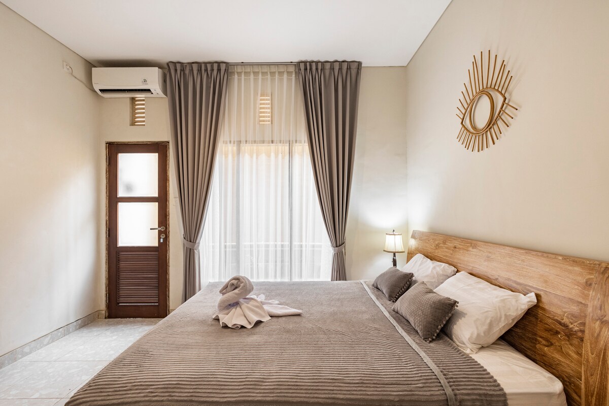 A bright sleeping area features a spacious bed with neutral-toned linens. Large windows adorned with sheer curtains allow natural light to fill the room. A wooden door and stylish round mirror add a touch of modern design to the inviting space.