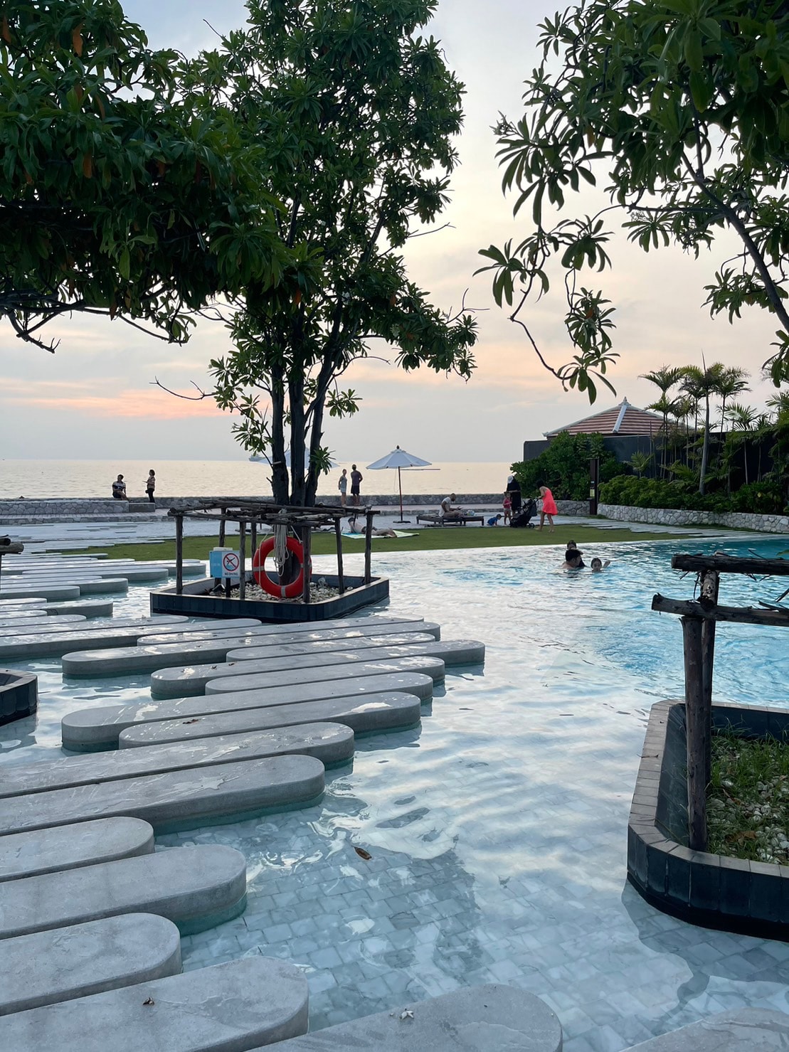 A scenic view of a swimming pool area is presented, featuring stepping stone pathways leading through clear water. Lush greenery surrounds the pool, and a distant beach can be seen at sunset. Guests enjoy leisurely activities nearby, adding a sense of relaxation to the atmosphere.