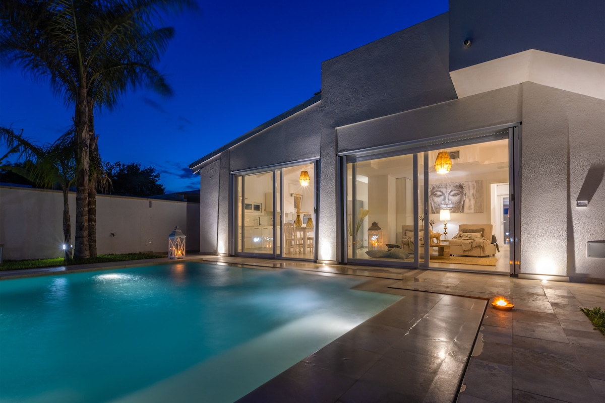 The exterior view of a modern villa is captured at night, showcasing a tranquil pool illuminated by soft lighting. Large glass doors provide an inviting glimpse into a cozy living area adorned with stylish furniture, while palm trees gently sway nearby.
