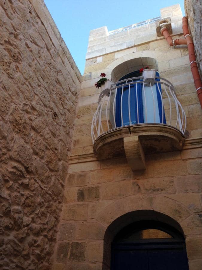 A charming balcony with white railing adorns the exterior of a traditional stone building. The balcony features potted flowers, adding a touch of color against the light-toned stone walls. A clear blue sky is visible above, enhancing the serene atmosphere.