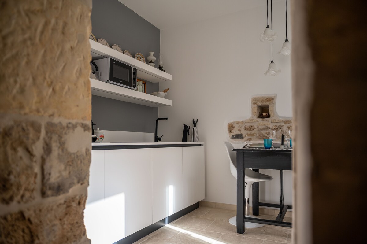 A modern kitchen is showcased, featuring sleek white cabinetry and open shelving displaying decorative plates. A dining table with a minimalist design sits adjacent to a vintage stone wall, and pendant lights provide soft illumination, enhancing the clean and functional layout.