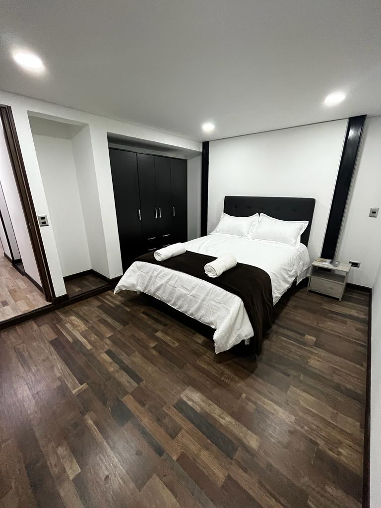 A well-lit bedroom features a spacious bed with crisp white linens and decorative towels. A dark wood wardrobe stands against the wall, and a small nightstand holds a lamp. The floor showcases rich, dark wood, contributing to a modern and uncluttered look.
