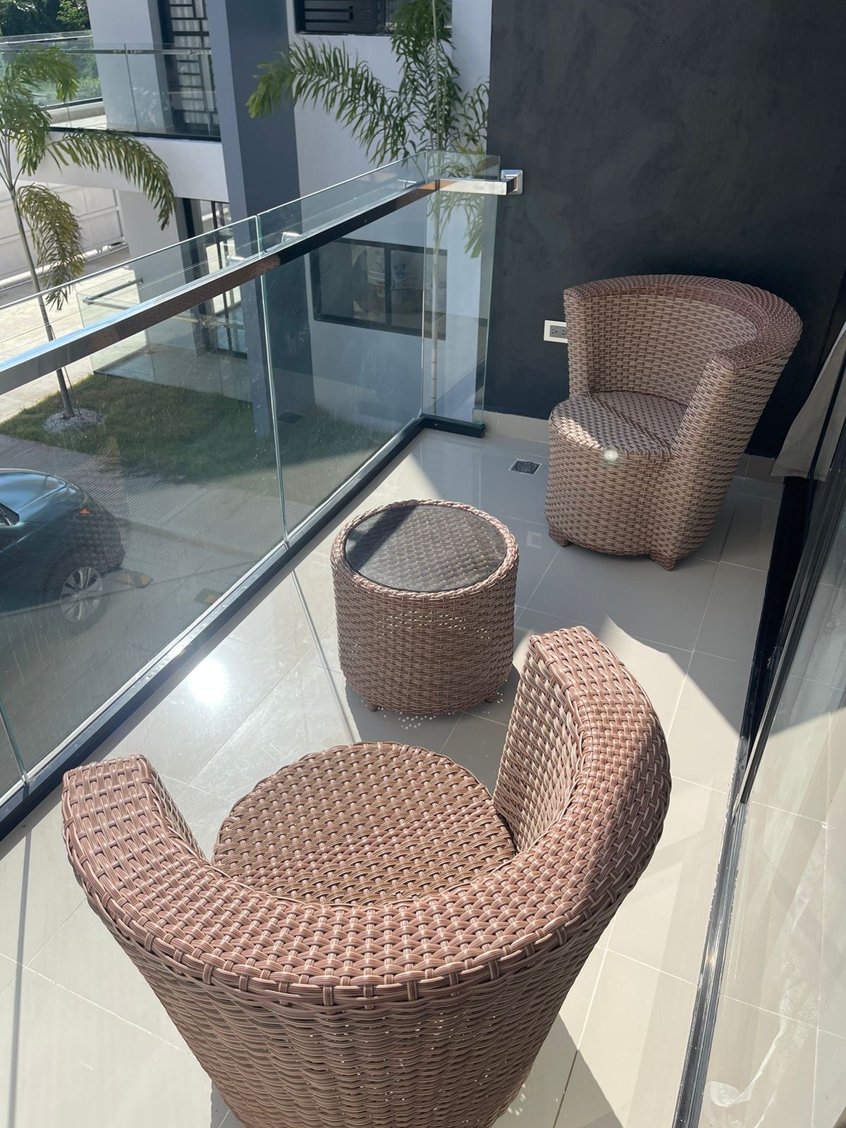 A balcony features two round, woven chairs and a matching round table. The glass railing provides unobstructed views of the surroundings. Sunlight reflects off the smooth tile flooring, enhancing the open and airy feel of the space.